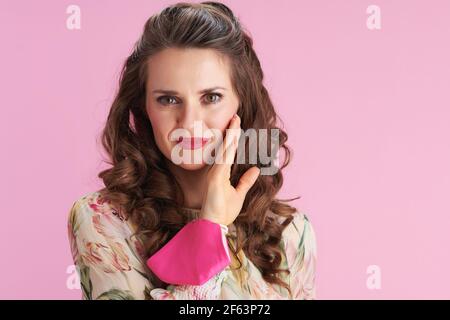 donna moderna in abito floreale con maschera rosa che controlla la pelle del viso isolato su sfondo rosa. Foto Stock