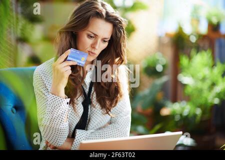 Green Home. Donna trendy con lunghi capelli ondulati con laptop e carta di credito seduta in una poltrona blu nel moderno soggiorno in giornata di sole. Foto Stock