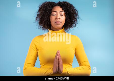 Donna africana calma che prega con le mani insieme simboleggia la preghiera e la gratitudine. Mudra. Concetto di yoga. Sfondo blu chiaro studio. Foto Stock