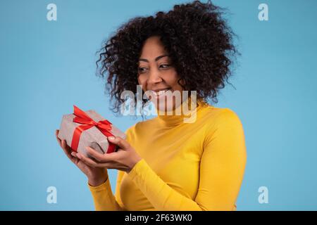 La donna africana eccitata ha ricevuto la scatola del regalo con l'arco. È felice e lusingata dall'attenzione. Ragazza sorridente con presente su sfondo blu. Studio Foto Stock