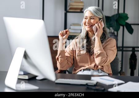 Sicuro di successo bella donna asiatica con capelli grigi senior, leader aziendale, manager, lavorare in ufficio, parlare al telefono con amici o colleghi, guardare via, sorridere Foto Stock