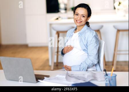 Gioiosa riuscita bella razza mista donna adulta incinta, freelancer, manager o business lady, lavorando a distanza, si siede alla scrivania, delicatamente tocca il suo ventre con le mani, guardare la fotocamera, sorridendo Foto Stock