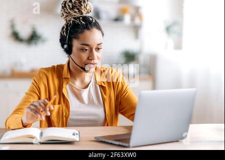 Donna afro-americana intelligente con dreadlock, in cuffia, in abiti casual, dipendente del call center, freelance o manager, guardando lo schermo del notebook, prendendo appunti Foto Stock