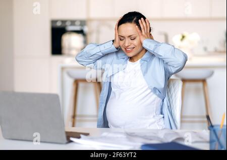 Una donna incinta, manager, designer o freelancer mista emaciata, irritata e stressata, che lavora a distanza, sperimentando stress e mal di testa, avendo bisogno di riposo, le mani che tengono chiuso gli occhi della testa Foto Stock