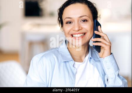 Conversazione telefonica. Primo piano di una donna da corsa mista felice e sicura vestita di camicia formale, segretaria, agente, manager, parlando con gli amici o colleghi su smartphone, guarda il sorriso amichevole della fotocamera Foto Stock