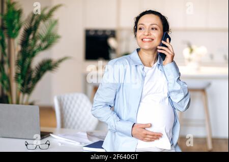 Gioioso successo bella donna incinta occupato, broker, manager o freelancer, lavorando a casa, parlando al telefono con un dipendente o cliente, in piedi vicino al tavolo, guardando via, sorridendo Foto Stock