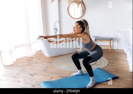 Stile di vita sano e sportivo. Attraente giovane afroamericana slim ragazza sportiva con Dreadlocks cura la sua salute, fa sport a casa su tappetino fitness, si siede-up con le braccia allungate, sorride Foto Stock