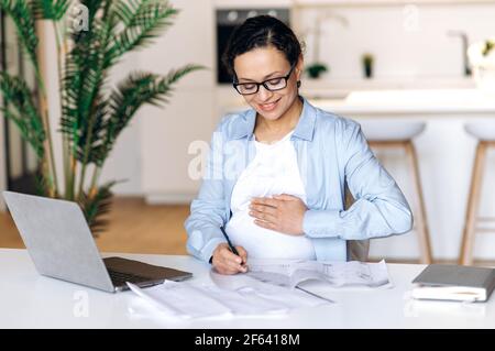 Donna incinta dipendente. Simpatica donna di gara mista incinta di successo, designer o top manager, lavora a casa da remoto, prendendo appunti e controllando il progetto aziendale, stracciando la pancia, sorridendo Foto Stock