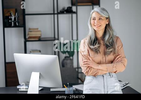 Ritratto di una donna asiatica con capelli grigi abbastanza influente, leader di affari, manager, ceo si trova in un ufficio moderno, elegantemente vestito, braccia incrociate, guardando al fianco, sorridente amichevole Foto Stock