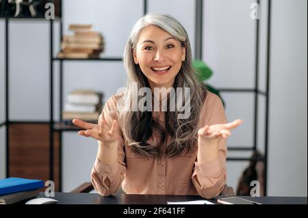Felice attraente donna di affari asiatica matura di successo, manager, seduto al tavolo, parlando in conferenza online, videochiamata con il team di affari o la famiglia, gesturing con le mani, sorridente Foto Stock