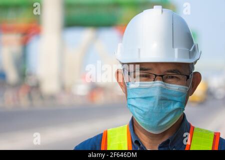 Primo piano uomo asiatico ingegnere indossare maschera medica lavorando nella costruzione del sito, vecchio uomo concetto età di pensionamento Foto Stock