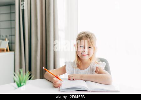 Una piccola ragazza sorridente si siede al tavolo e scrive in un taccuino bianco. Concetto di istruzione. Scuola domestica. Compiti. Faccia sorridente. Foto di alta qualità Foto Stock