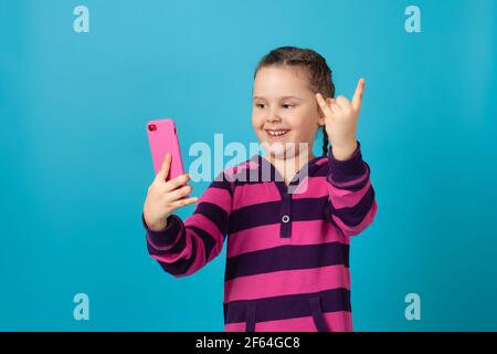 primo piano ritratto di una ragazza ridente con i pigtail che parlano in videochiamata, tenendo il telefono a braccio e mostrando segni di corna con le dita Foto Stock