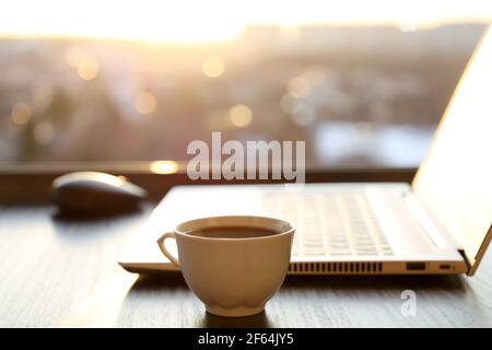 Ambiente di lavoro accogliente in ufficio a casa con una tazza di caffè caldo e un computer portatile sul tavolo contro la finestra al tramonto. Concetto di lavoro remoto e guadagnare online