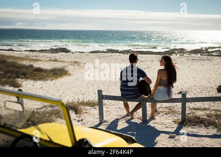 Felice coppia caucasica seduta sulla spiaggia in riva al mare giocando chitarra Foto Stock