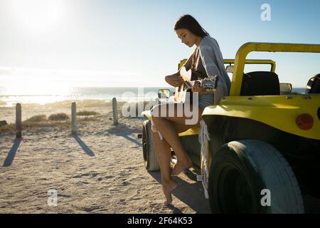 Donna caucasica felice seduta sulla spiaggia buggy vicino al mare suonare la chitarra Foto Stock