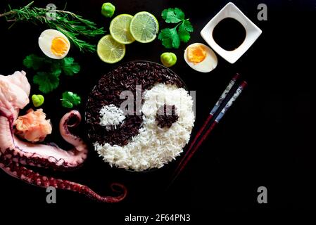 Una vista dall'alto di una ciotola di riso yin yang, chopsticks, fette di lime, uova bollite, salsa di soia, spezie, e tentacoli calamari su sfondo nero Foto Stock