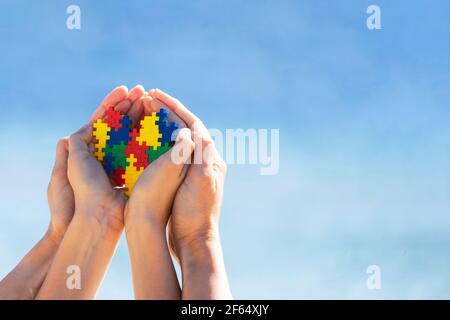 Mani autistiche di capretto con cuore di puzzle multicolore supportato dalle mani della madre su sfondo blu del cielo. Giornata mondiale di consapevolezza dell'autismo Foto Stock