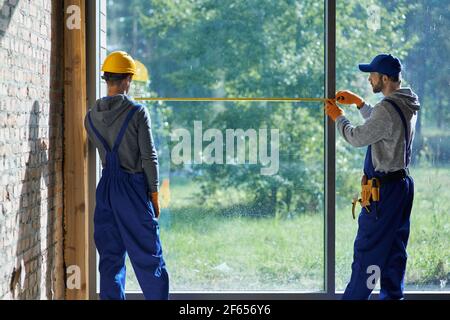 Fare nuova casa. Vista posteriore di due giovani costruttori maschili che indossano tute blu utilizzando un nastro di misurazione mentre si lavora sul cantiere Foto Stock