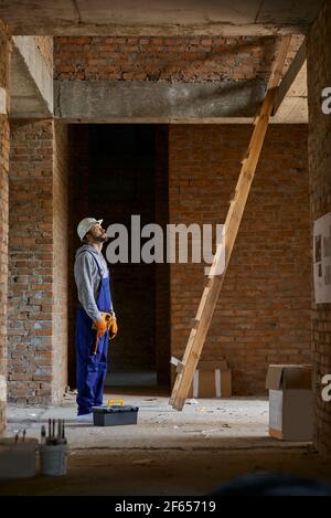 Foto a tutta lunghezza di giovane lavoratore in tute blu e elmetto guardando in alto mentre si va a salire la scala, lavorando sulla costruzione della casa Foto Stock