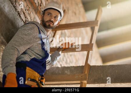 Ritratto di bel costruttore maschile in tute blu e elmetto che guarda via, salendo lungo la scala mentre si lavora in cantiere Foto Stock