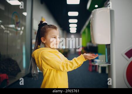 Bambina che usa l'erogatore automatico di gel di alcool spruzzando sulle mani disinfettante disinfettante antisettico macchina igienizzante, nuova vita normale dopo la pandemia di Coronavirus COVID-19. Foto Stock