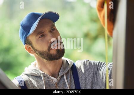 Primo piano ritratto di un giovane costruttore con cappuccio blu e tute utilizzando il nastro di misurazione mentre si lavora sulla costruzione di cottage in una giornata di sole Foto Stock