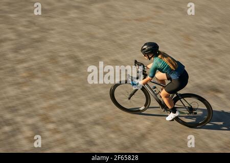 Inquadratura ad alto angolo di una ciclista femminile professionista con indumento da ciclismo e attrezzatura protettiva che si concentra durante la guida in bicicletta, allenandosi all'aperto Foto Stock