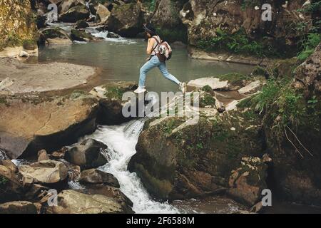 Elegante donna viaggiatore con zaino camminare sulle rocce del fiume in montagna. Giovane donna in stoffa casual e cappello che salta attraverso il fiume. Viaggi e wa Foto Stock