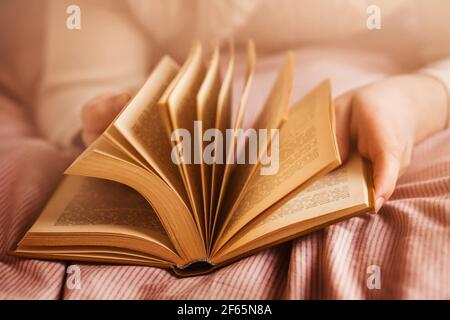 Un uomo in un maglione bianco giace su un letto con una coperta rosa e tiene un interessante vecchio libro tra le sue mani, che si capovolge attraverso le sue pagine alla luce del sole. Foto Stock