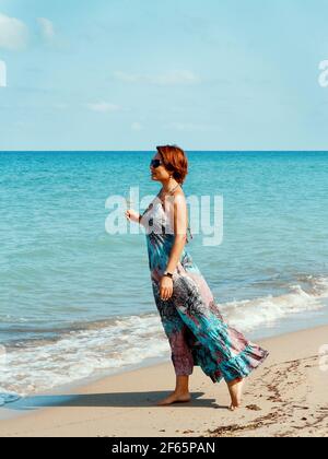 Giovane caucasica in un lungo vestito con bicchiere di vino in mano a piedi sulla spiaggia Foto Stock