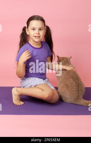 Ritratto a lunghezza intera di una ragazza con coda di pony in una T-shirt viola e shorts che tengono un gatto rosso su un tappeto di sport viola, isolato su sfondo rosa Foto Stock