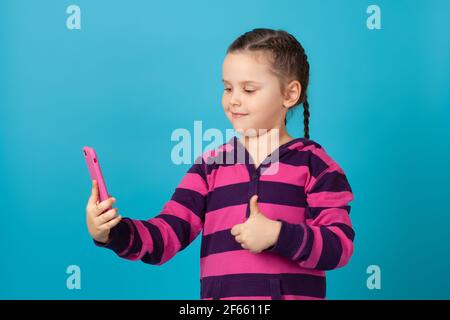 primo piano ritratto di una ragazza di cinque anni con i pigtail che parlano su una connessione di videotelefono e che danno un pollice in su, isolato su uno sfondo blu Foto Stock