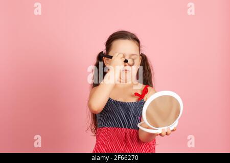 divertente, goffa ragazza di cinque anni segue la madre e cerca di fare con un pennello in polvere isolato su uno sfondo rosa Foto Stock