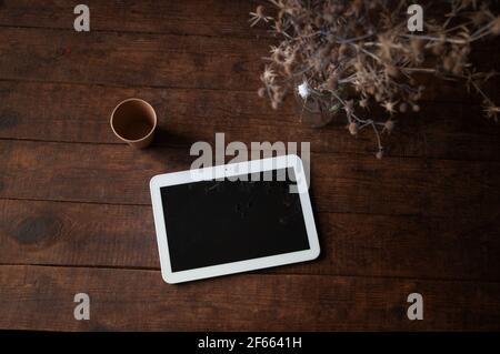 Ambiente di lavoro moderno con tablet pc sul tavolo. Sul tavolo c'è un computer tablet con fiori secchi in un bicchiere e una tazza di caffè usa e getta Foto Stock