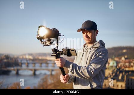 Giovane uomo che riprende con macchina fotografica e gimbal. Regista in piedi contro lo skyline urbano. Praga, Repubblica Ceca Foto Stock