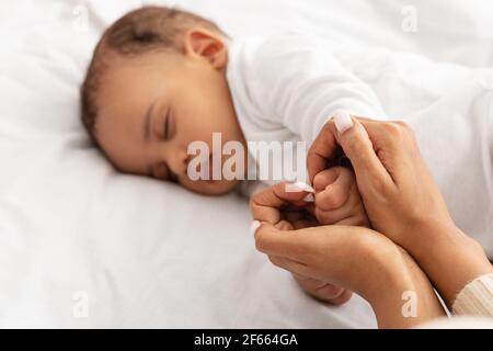 Amorevole mamma nera che tiene addormentata la mano del bambino indoor, fuoco selettivo Foto Stock