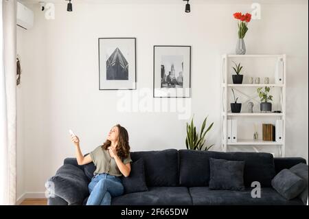 La donna si siede sul divano in soggiorno e si sente calda, tiene il commutatore di controllo remoto e accende il condizionatore d'aria, fugge dal calore estivo a casa sotto un flusso di aria fredda da un sistema separato Foto Stock