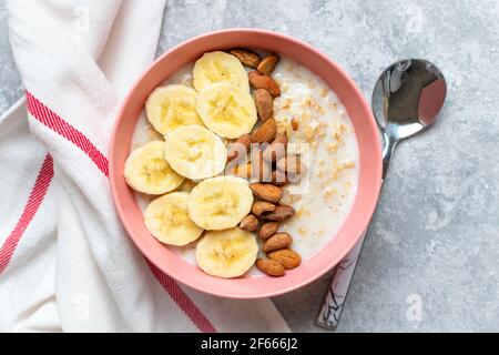 Farinata d'avena con banana, mandorle noci in ciotola rosa, bicchiere di latte, tovagliolo su fondo grigio in cemento Vista dall'alto colazione o pranzo sani ingredienti naturali Foto Stock