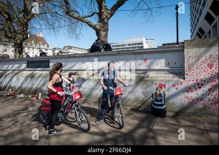 Londra, Regno Unito. 30 Marzo 2021. I ciclisti passano un volontario che attingono i cuori su un muro a Lambeth, vicino al Tamigi, con ogni cuore che rappresenta qualcuno che è morto durante la pandemia di coronavirus in corso nel Regno Unito. Chiamato il National Covid Memorial Wall, è stato creato dalle famiglie covide-19 in lutto per la giustizia e si estende per mezzo miglio entro il momento in cui è completo. Credit: Stephen Chung/Alamy Live News Foto Stock