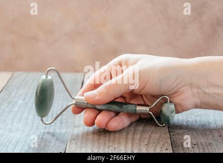 Mano che tiene un rullo di massaggio guasha di giada verde utilizzato per il massaggio del viso su sfondo di legno, SPA relax, viso e cura della pelle sfondo Foto Stock