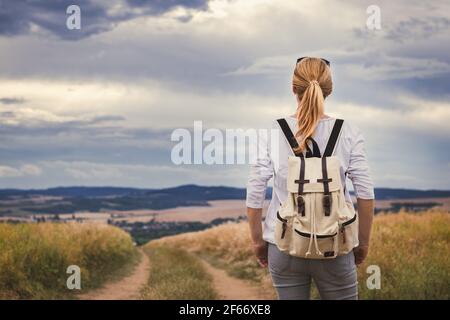 Donna con zaino che gode di vista su un bellissimo paesaggio con cielo drammatico. Turista femminile godendo di viaggio in campagna in estate Foto Stock