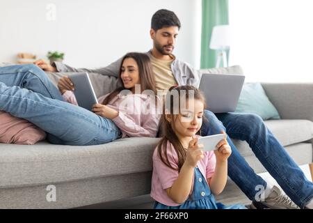 Dipendenza da gadget. Famiglia orientale che utilizza diversi dispositivi elettronici, che si riposano insieme in salotto a casa Foto Stock