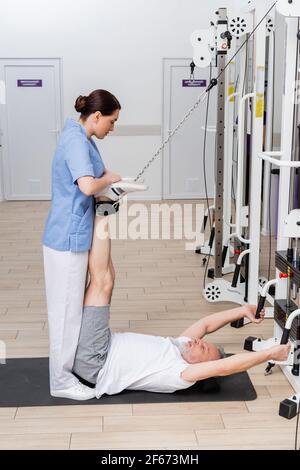 brunette fisioterapista che sostiene le gambe di un uomo maturo che si esercita in forma fisica mat Foto Stock