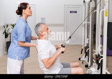 vista laterale del riabilitologo che sostiene l'uomo maturo che esercita sulla trazione macchina per cavi Foto Stock