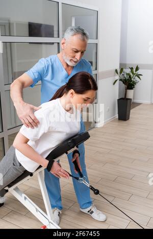 allenatore di media età che assiste la donna bruna durante la riabilitazione fisica Foto Stock