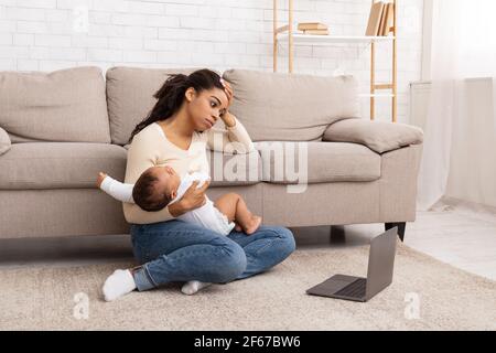 Esausta madre nera che tiene piangendo bambino seduto al laptop indoor Foto Stock