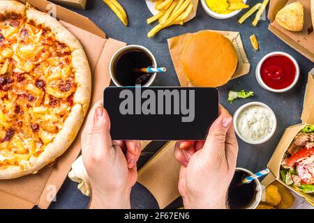 Consegna fastfood ordinare online concetto di cibo. Grande set di cibi assortiti da take out, mani con smartphone in vista pic flatlay Rop Foto Stock