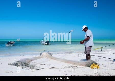 Un pescatore sulla spiaggia di Holbox Island in Messico ripara le sue reti sulla spiaggia la mattina presto. Sullo sfondo il Mar dei Caraibi e piccolo Foto Stock