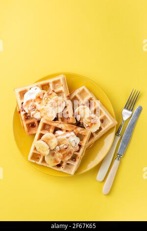 Waffle con banana, fette di mandorle, crema e salsa al caramello isolati su sfondo giallo, vista dall'alto. Colazione dolce o waffle da dessert Foto Stock